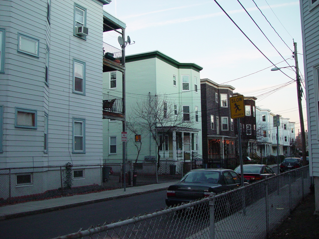 Wooden triple-decker houses, stacked porches and parked cars on a residential street in Cambridge.